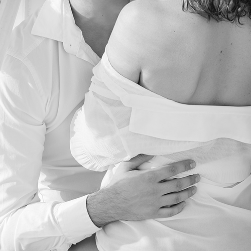 Couple standing closely in loose white shirts, the man has his hand on the woman's waist